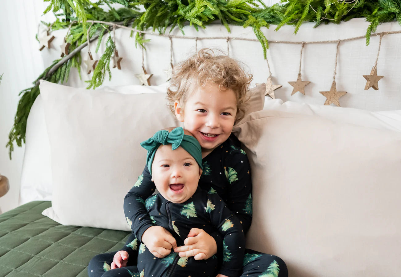 Two children in matching pajamas sitting on a bed with green bedding and Christmas decorations.